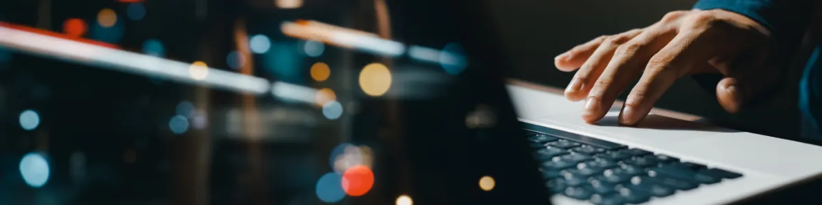 Close‑up of a hand typing on a laptop keyboard with blurred lights reflected across the screen.