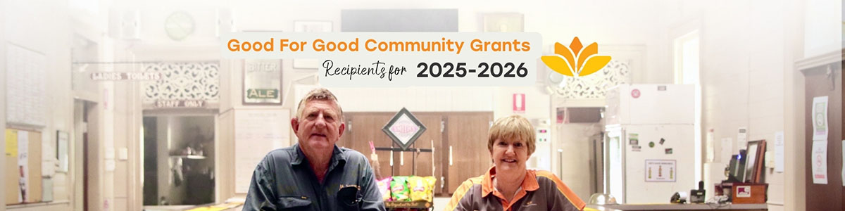 Two people seated at a table in a community hall with signage announcing Good For Good Community Grants Recipients for 2025-2026 and the Queensland Country Bank logo above them.