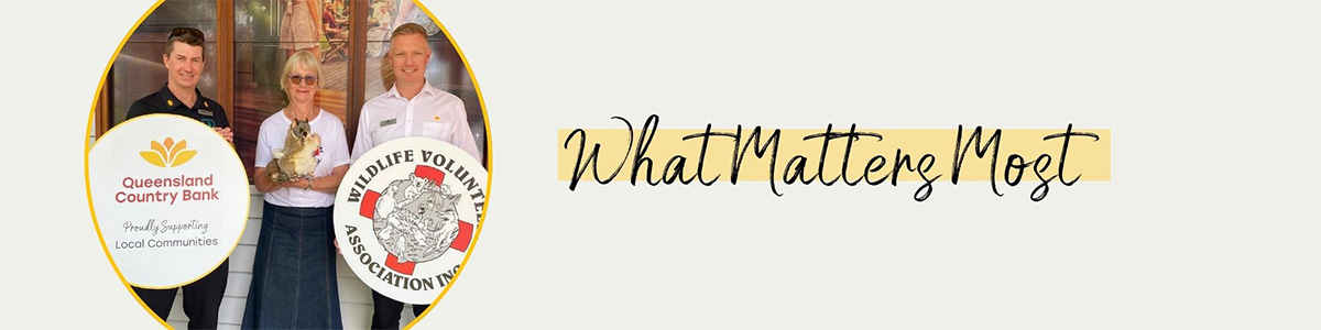 Three people standing indoors holding an animal, with two circular logos in the foreground. Left logo reads 'Queensland Country Bank Proudly Supporting Local Communities' with a yellow flower icon. Right logo reads 'Wildlife Volunteers Association Inc.' featuring an illustration of animals. Text on the right says 'What Matters Most' in black script on a light background.