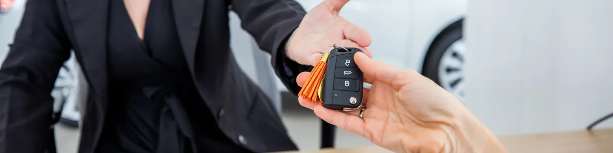 Close-up of a car key handover across a desk, with documents, a keyboard, and a computer mouse visible, and a vehicle in the background.