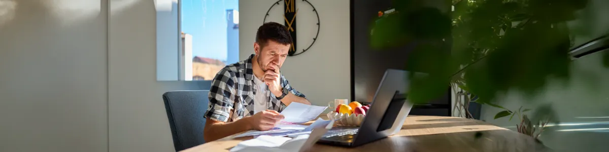Person sitting at a table reviewing bills and documents beside a laptop, with papers spread out, illustrating financial stress and budgeting at home.